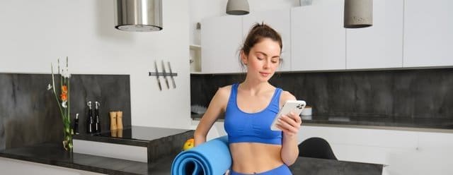 Portrait of attractive, fit young woman in sportswear, standing in living room with rubber yoga mat, holding smartphone, doing fitness exercises with mobile app at home