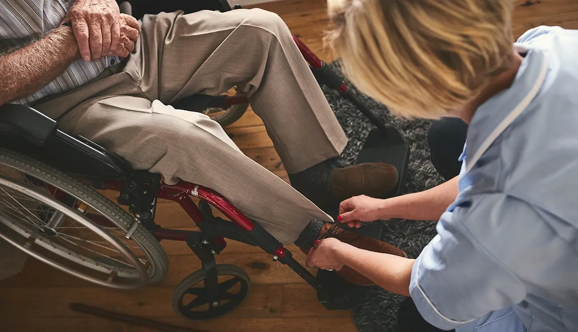 Caregiver assists a person in a wheelchair by fastening adaptive clothing shoes for easier dressing.